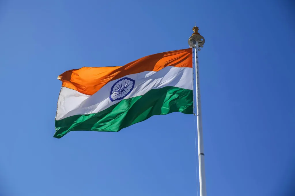 The national flag of India, featuring horizontal stripes of saffron, white, and green with a navy blue Ashoka Chakra in the center, waving on a flagpole against a clear blue sky